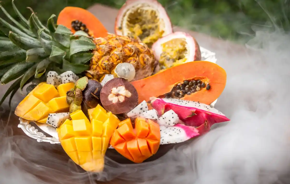 A tray filled with exotic tropical fruits including pineapple, papaya, mango, passion fruit, dragon fruit, and mangosteen, surrounded by smoke.