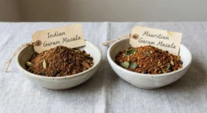 Side-by-side ceramic bowls, one labeled "Indian Garam Masala" and the other "Mauritian Garam Masala" containing a distinct Mauritius masala UK blend with visible whole spices and curry leaves.