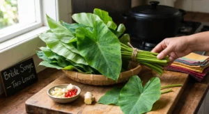 A hand reaching for a fresh bundle of Brède Songe (taro leaves) in a woven basket on a kitchen counter, next to a chalkboard sign, garlic, ginger, and red chilies.